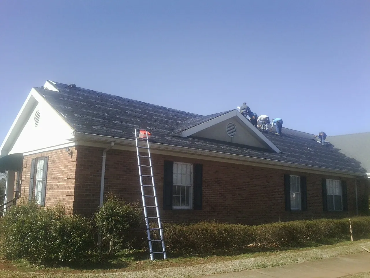 Crew tearing shingles off a brick ranch roof with a ladder set — Metal Roof Repair work by our crew in Stokes