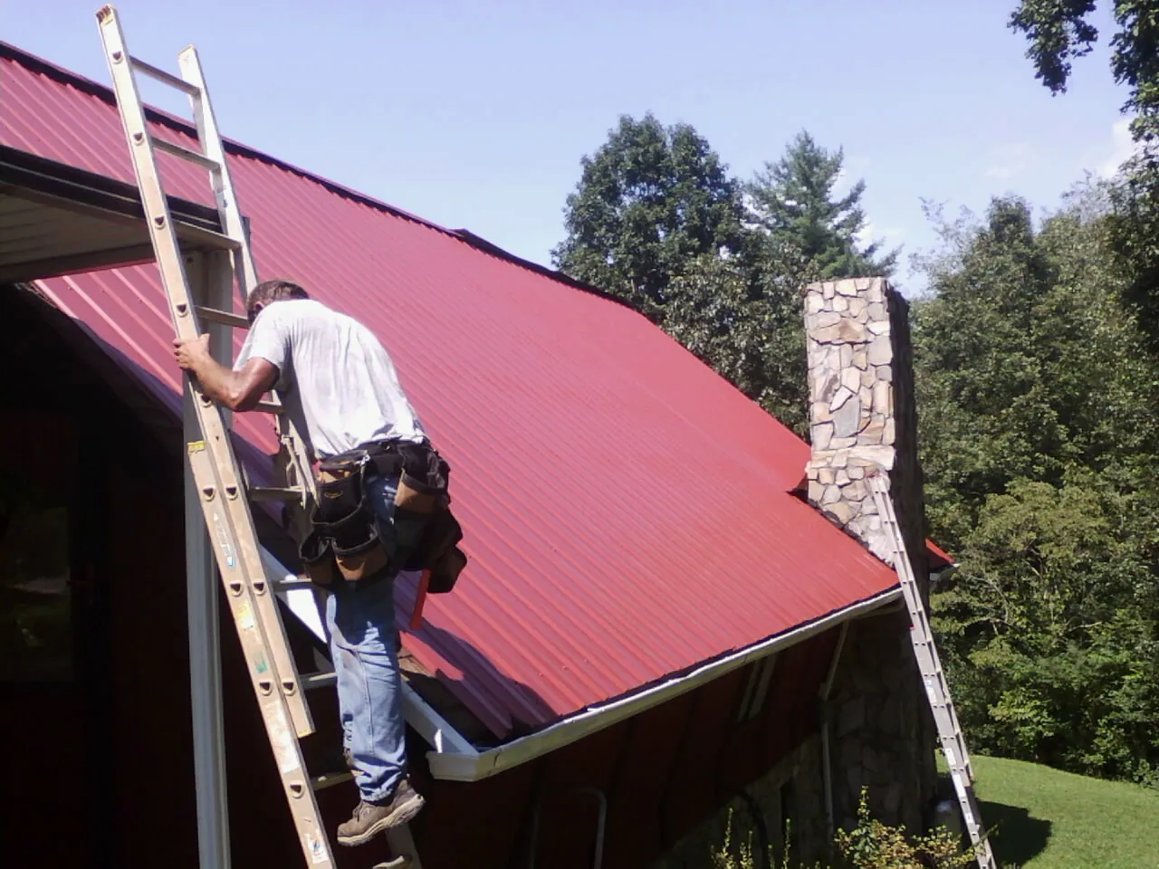 Roofer on a ladder beside a red metal roof and stone chimney — Soffit & Fascia Repair by Roofing Brothers in Stokes